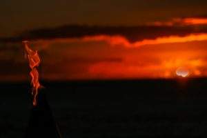 Sunset over ocean in background with torch flame burning straight up on left side of image