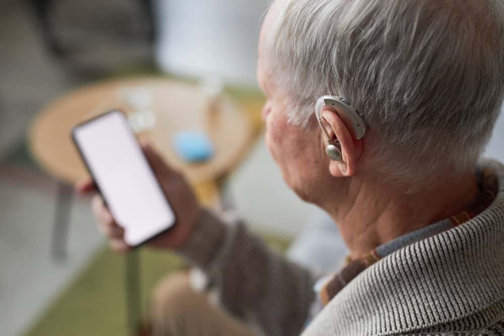 Older adult using a hearing aid while looking at a phone, illustrating VA hearing loss rating and hearing impairment evaluation.
