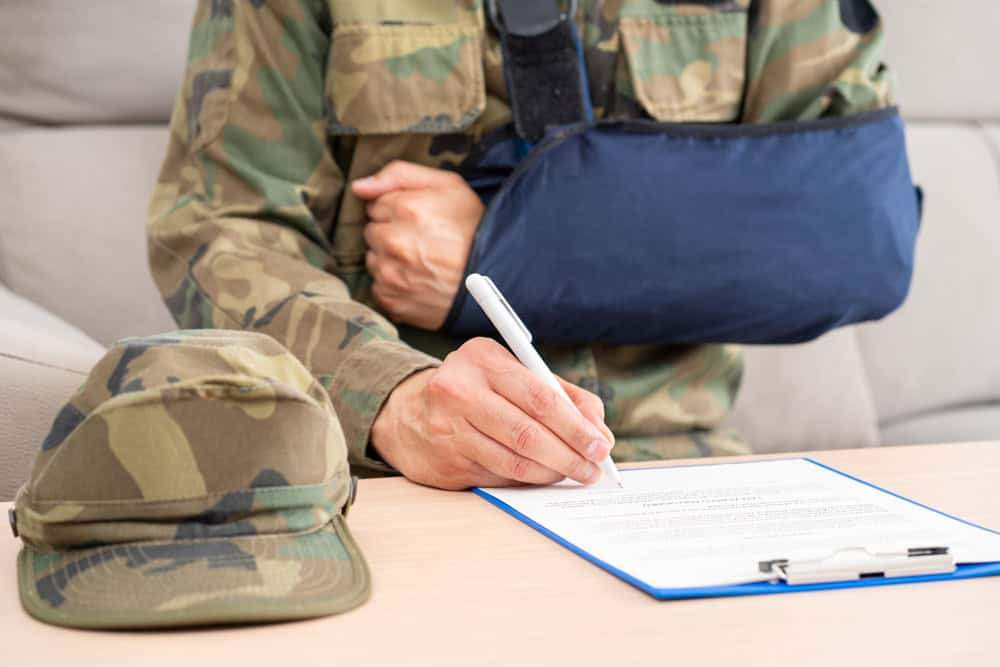A person in a military uniform with their arm in a dark blue sling, signing a document on a clipboard. A camouflage cap is on the table.